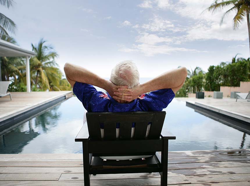 Older man with gray hair sitting in lounge chair by pool with arms behind head relaxing